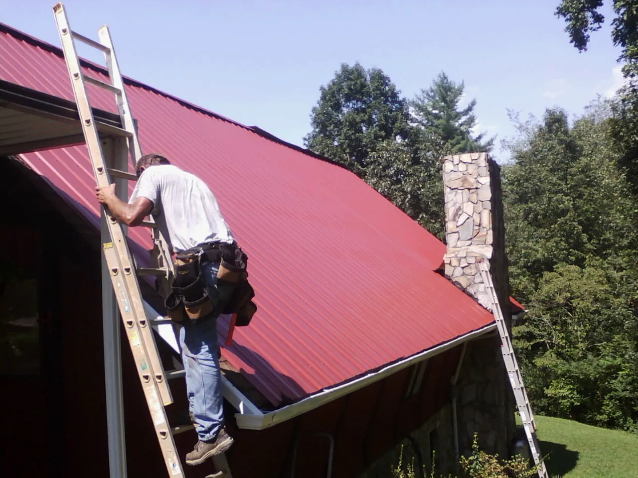 Roofer on a ladder beside a red metal roof and stone chimney — Soffit & Fascia Repair by Roofing Brothers in Rimforest